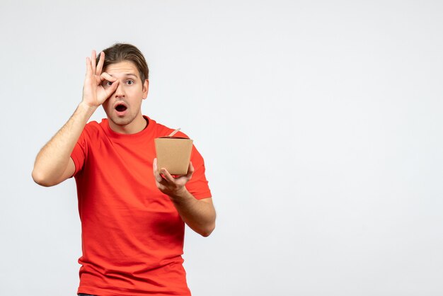 Vista frontal del joven sorprendido en blusa roja sosteniendo una pequeña caja haciendo gesto de anteojos sobre fondo blanco.