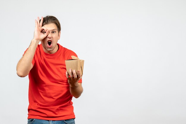 Vista frontal del joven sorprendido en blusa roja sosteniendo una pequeña caja haciendo gesto de anteojos sobre fondo blanco.