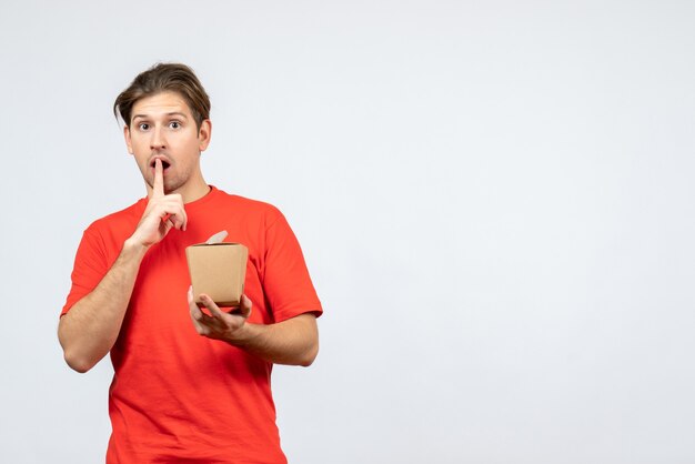 Vista frontal del joven sorprendido en blusa roja sosteniendo una pequeña caja haciendo gesto de silencio sobre fondo blanco.