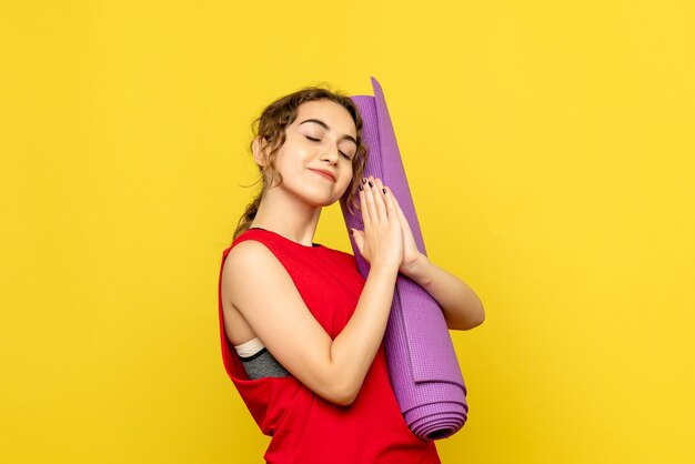 Vista frontal de la joven sosteniendo una alfombra púrpura en la pared amarilla