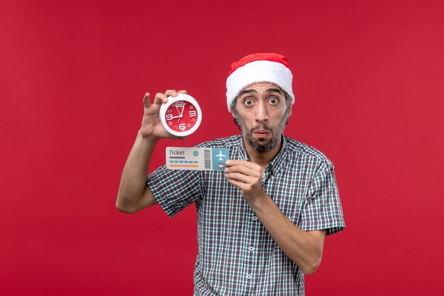 Vista frontal joven sosteniendo boleto y reloj en pared roja tiempo de emoción masculina roja