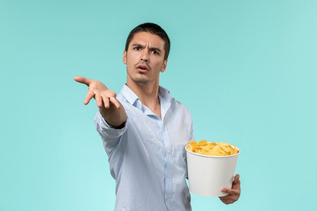 Vista frontal joven sosteniendo una canasta con patatas cips comiendo y viendo películas en la pared azul cine de películas remotas y solitarias