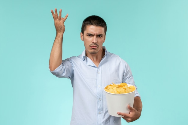 Vista frontal joven sosteniendo una canasta con patatas cips comiendo y viendo películas en la pared azul claro cine de películas remotas y solitarias