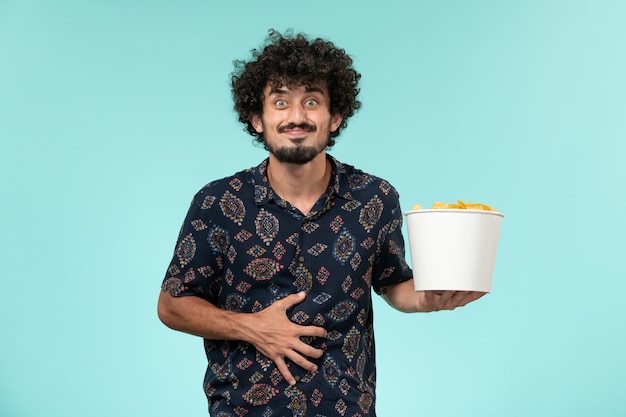 Vista frontal joven sosteniendo la canasta con patatas cips en una pared azul control remoto de película de cine masculino