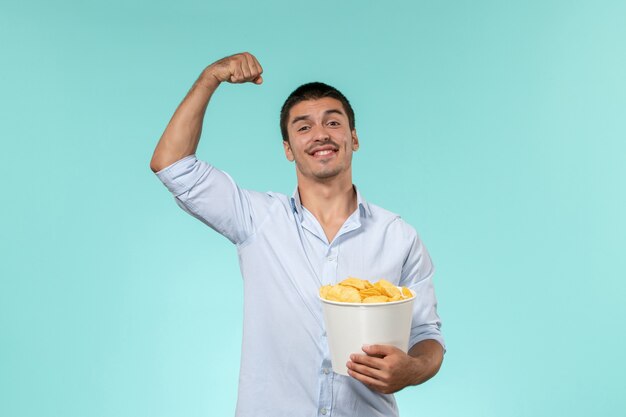 Vista frontal joven sosteniendo cips de patata y flexionando en una pared azul cine masculino remoto solitario