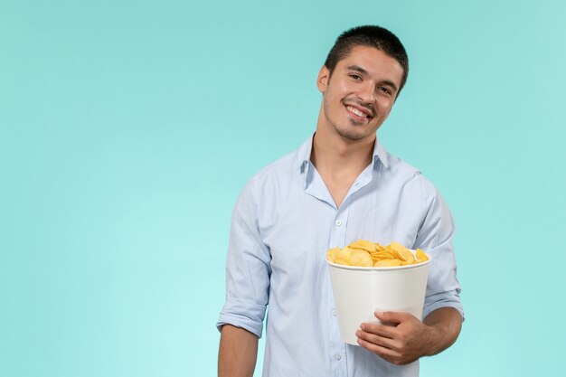 Vista frontal joven sosteniendo cips de patata y sonriendo en una pared azul cine masculino remoto solitario