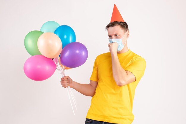 Vista frontal del joven sosteniendo globos de colores en máscara estéril tosiendo en la pared blanca
