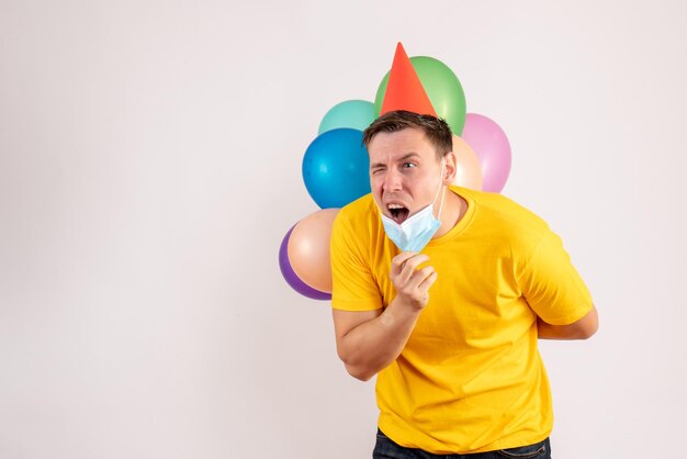 Vista frontal del joven sosteniendo globos de colores en máscara en la pared blanca