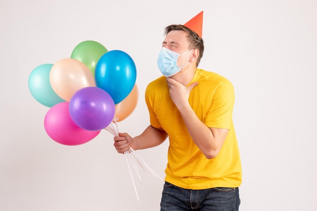 Vista frontal del joven sosteniendo globos de colores en la máscara que tiene problemas para respirar en la pared blanca