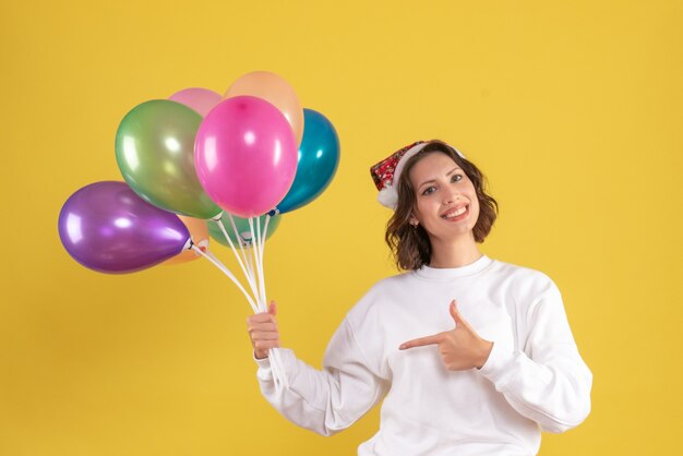 Vista frontal de la joven sosteniendo globos de colores en la pared amarilla