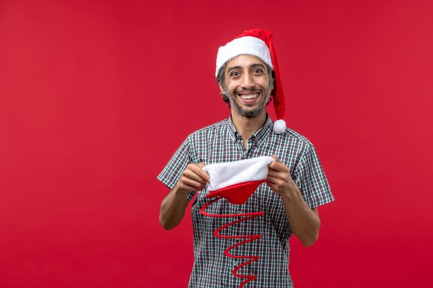 Vista frontal del joven sosteniendo la gorra roja del juguete de Navidad en la pared roja