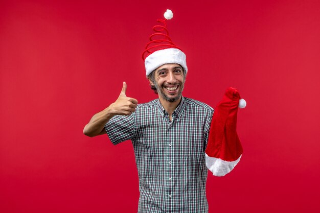 Vista frontal del joven sosteniendo gorro de navidad en la pared roja