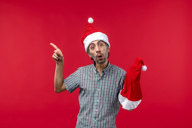 Vista frontal del joven sosteniendo gorro rojo de Navidad en la pared roja