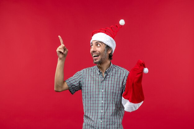 Vista frontal del joven sosteniendo gorro rojo de Navidad en la pared roja