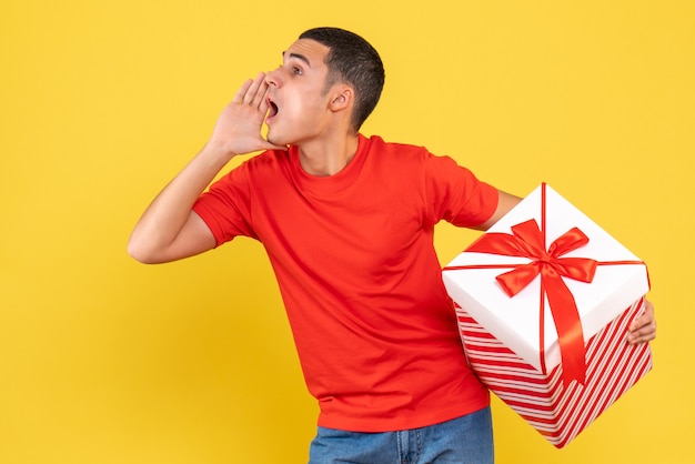 Vista frontal del joven sosteniendo un gran regalo de Navidad llamando a alguien en la pared amarilla