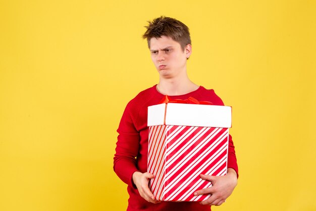Vista frontal del joven sosteniendo gran regalo de Navidad en la pared amarilla