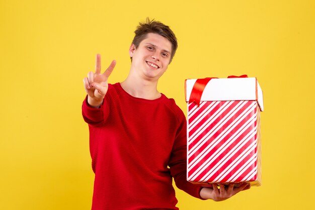 Vista frontal del joven sosteniendo gran regalo de Navidad sonriendo en la pared amarilla