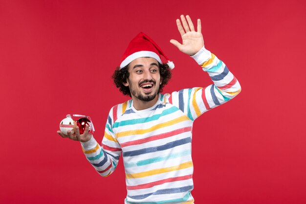 Vista frontal joven sosteniendo juguetes de árbol de navidad en la pared roja vacaciones año nuevo humano rojo