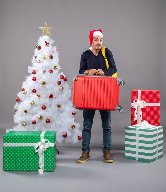Vista frontal joven sosteniendo la maleta y la mochila de pie cerca del árbol de Navidad en gris aislado