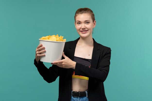 Vista frontal joven sosteniendo el paquete con papas fritas sonriendo y viendo la película en la superficie azul