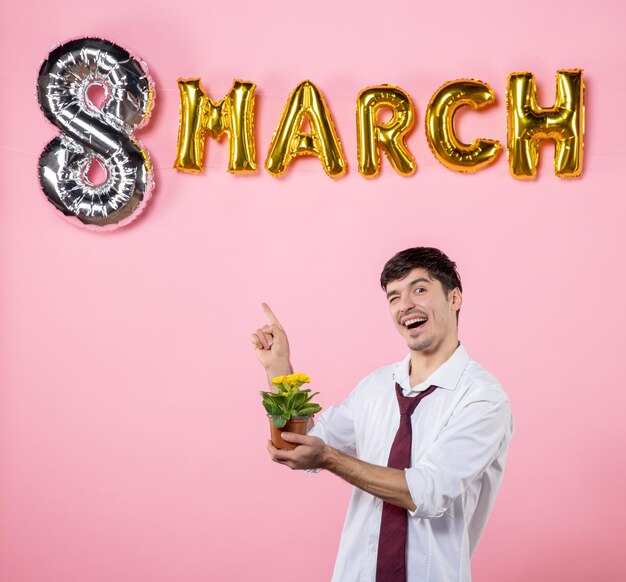 Vista frontal joven sosteniendo una pequeña flor en una maceta con decoración de marzo sobre fondo rosa hombre color día de la mujer matrimonio igualdad presente fiesta femenina