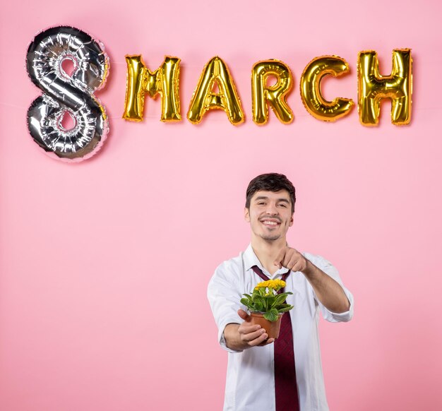 Vista frontal joven sosteniendo una pequeña flor en una maceta con decoración de marzo sobre fondo rosa presente hombre matrimonio igualdad día de la mujer color fiesta femenino