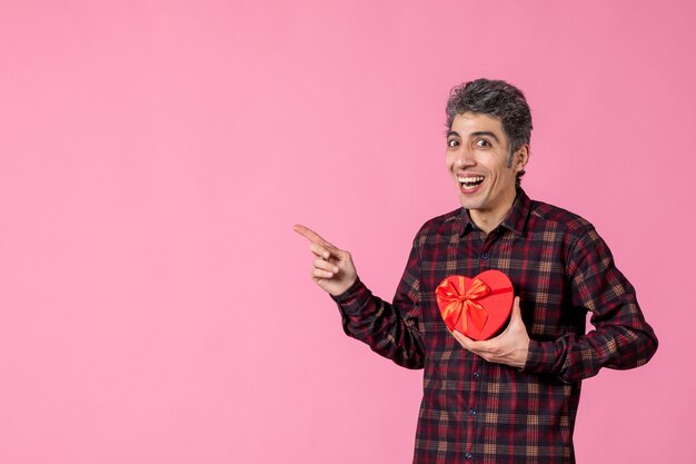 Foto gratuita vista frontal joven sosteniendo presente en forma de corazón rojo en la pared rosa