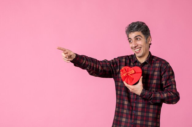 Vista frontal joven sosteniendo presente en forma de corazón rojo en la pared rosa