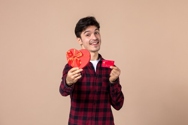 Vista frontal joven sosteniendo presente en forma de corazón y tarjeta bancaria en la pared marrón