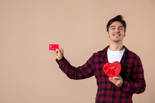 Vista frontal joven sosteniendo presente en forma de corazón y tarjeta bancaria en la pared marrón