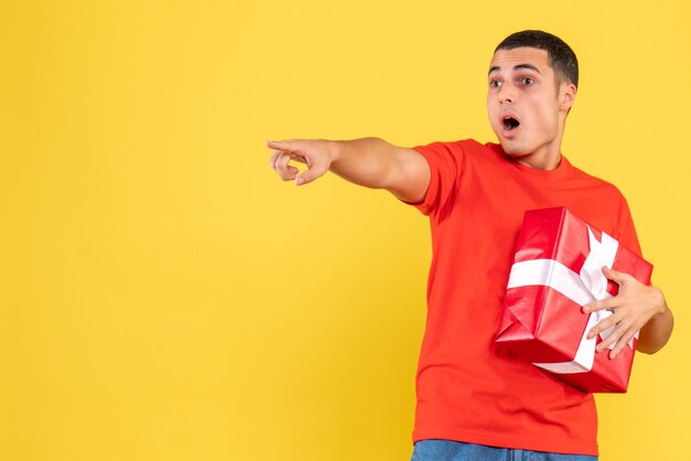 Vista frontal del joven sosteniendo el presente sorprendentemente apuntando a la pared amarilla