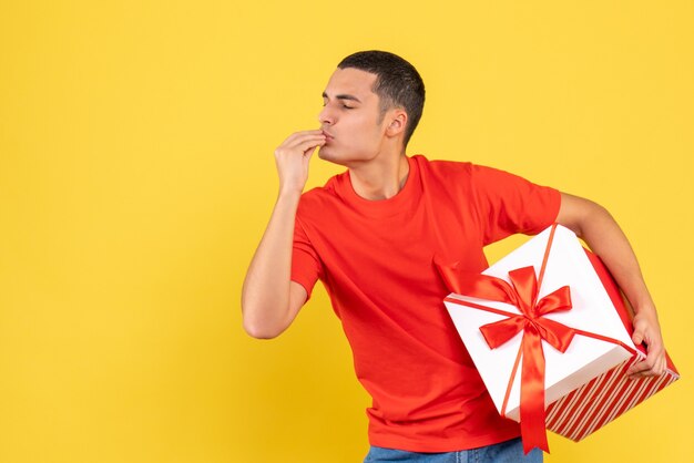 Vista frontal del joven sosteniendo un regalo de Navidad en una pared amarilla