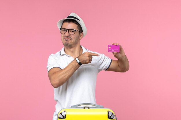 Vista frontal del joven sosteniendo una tarjeta bancaria púrpura en vacaciones de verano en la pared rosa