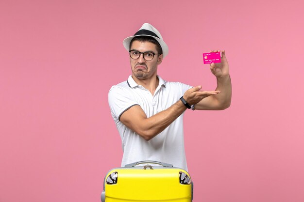 Vista frontal del joven sosteniendo una tarjeta bancaria de vacaciones en la pared rosa