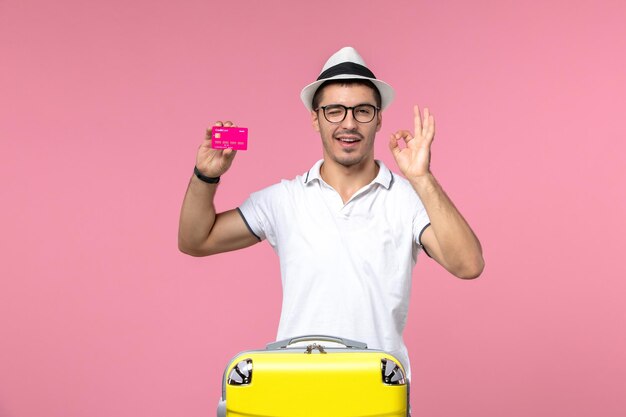 Vista frontal del joven sosteniendo una tarjeta bancaria de vacaciones en la pared rosa