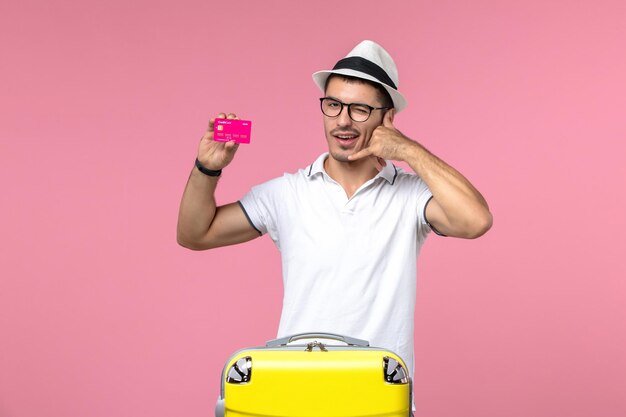 Vista frontal del joven sosteniendo una tarjeta bancaria de vacaciones en la pared rosa