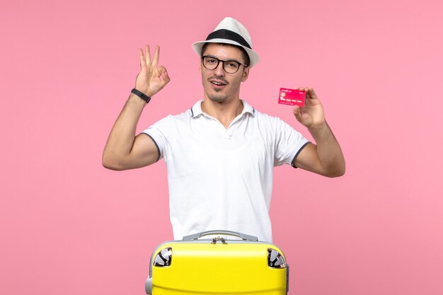 Vista frontal del joven sosteniendo una tarjeta bancaria de vacaciones en la pared rosa