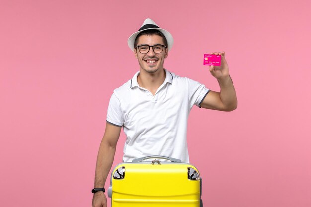 Vista frontal del joven sosteniendo una tarjeta bancaria en las vacaciones de verano sonriendo en la pared rosa