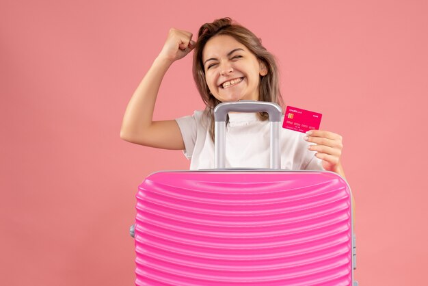 Vista frontal del joven sosteniendo la tarjeta detrás de la maleta rosa en la pared rosa