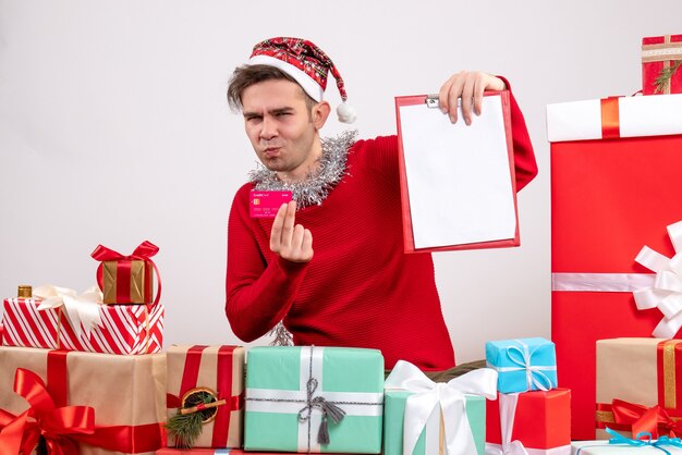 Vista frontal joven sosteniendo la tarjeta y el portapapeles sentado alrededor de los regalos de Navidad