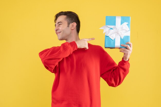 Vista frontal joven con suéter rojo apuntando a caja de regalo sobre fondo amarillo