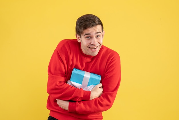 Vista frontal joven con suéter rojo sosteniendo su regalo apretado sobre fondo amarillo