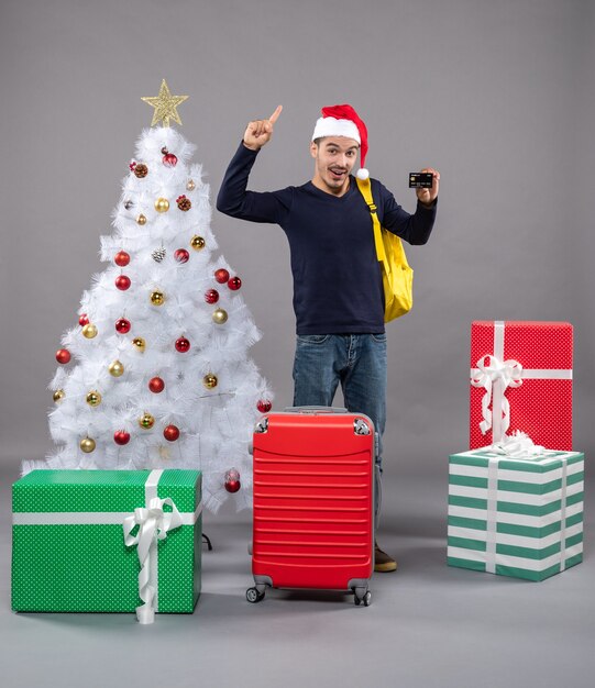 Vista frontal joven con tarjeta y maleta roja cerca del árbol de Navidad y presenta en gris aislado