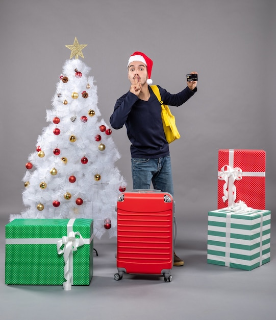 Vista frontal joven con tarjeta y maleta roja haciendo cartel de shh de pie cerca del árbol de Navidad en gris aislado