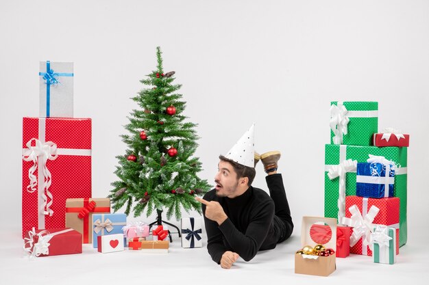 Vista frontal del joven tendido alrededor de regalos de vacaciones en la pared blanca