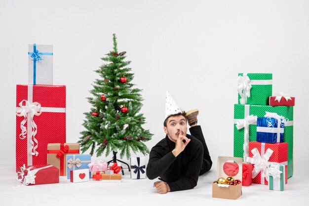 Vista frontal del joven tendido alrededor de regalos de vacaciones en la pared blanca