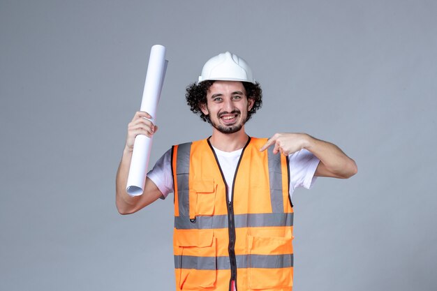 Vista frontal del joven trabajador de la construcción confiado en chaleco de advertencia con casco de seguridad y mostrando en blanco apuntando a sí mismo en la pared gris