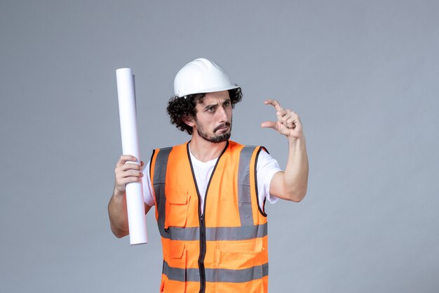 Vista frontal del joven trabajador de la construcción masculino preocupado en chaleco de advertencia con casco de seguridad y mostrando en blanco en la pared gris