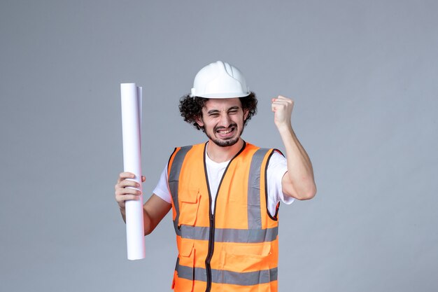Vista frontal del joven trabajador de la construcción sonriente feliz en chaleco de advertencia con casco de seguridad y mostrando en blanco en la pared gris