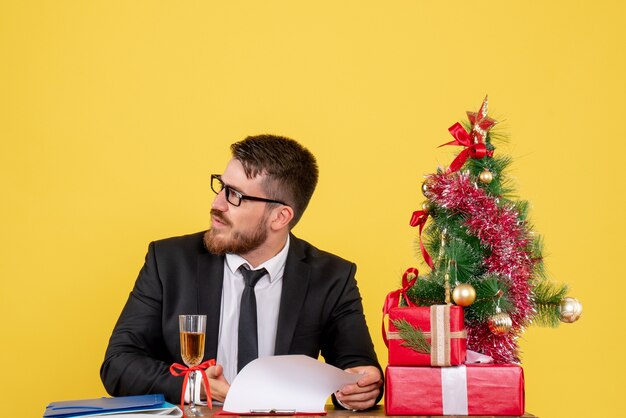 Vista frontal joven trabajador detrás de su mesa con regalos y árbol de Navidad en amarillo